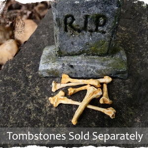 May include: A small, gray tombstone with the inscription "RIP" sits on a gray, textured surface.  A pile of small, yellow, plastic bones are scattered in front of the tombstone.  Tombstones sold separately.