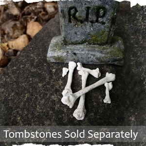 May include: White plastic bones arranged in a cross shape on a grey stone surface. A small grey tombstone with the inscription "RIP" is in the background. Tombstones sold separately.