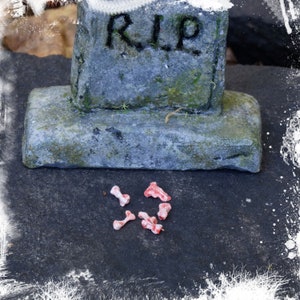 Peut inclure: Une pierre tombale grise et usée avec l'inscription "RIP" repose sur une surface gris foncé. Plusieurs petits os en plastique blanc et rouge sont éparpillés autour de la base de la pierre tombale.