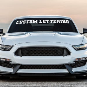 May include: A white sports car with a black grill and the text "CUSTOM LETTERING" across the top of the windshield. The car is parked outside on a cloudy day.