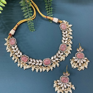 Puede incluir: Un conjunto de collar y pendientes dorados con piedras rosas cuadradas y detalles de perlas blancas. El collar y los pendientes tienen un diseño a juego, con un delicado patrón de hojas y un cordón dorado.