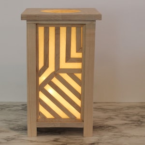Puede incluir: Una lámpara de madera con un patrón geométrico recortado en los lados. La lámpara está encendida y proyecta un brillo cálido.