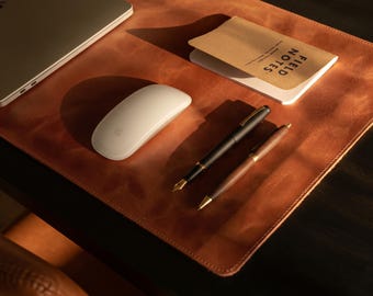 May include: A tan leather desk mat featuring a white computer mouse, a Field Notes notebook, and two pens, one black and one brown, arranged on the surface alongside a portion of a silver laptop.