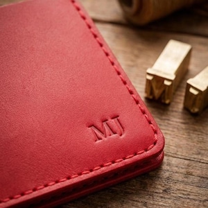 May include: A close-up of a red leather wallet with the initials "MJ" embossed on the corner. The wallet is stitched with a contrasting thread. A spool of thread and metal letter stamps are in the background.