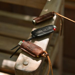 May include: Three leather lighter cases in varying shades of brown and black, each with a lighter inside. The cases have decorative stitching and are attached to leather cords. The lighters have red ignition buttons.