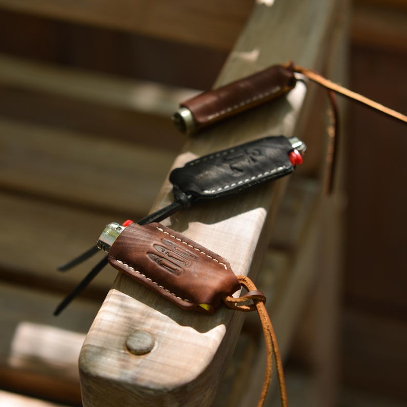 May include: Three leather lighter cases in varying shades of brown and black, each with a lighter inside. The cases have decorative stitching and are attached to leather cords. The lighters have red ignition buttons.