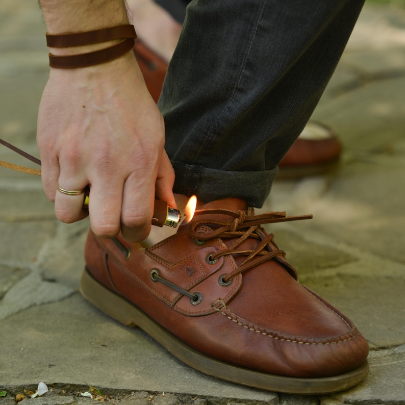 May include: Brown leather boat shoes with brown laces. A person is holding a lighter near the laces. A brown leather bracelet is wrapped around the wrist.