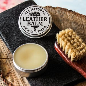 May include: A silver tin of leather balm with the text "ALL NATURAL LEATHER BALM MADE IN UKRAINE". An open tin of balm and a wooden-handled brush are on a dark grey cloth and wooden surface.