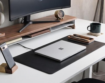 May include: A dark wood monitor stand sits on a white desk, supporting a black computer monitor. A silver laptop rests on a black desk mat. A wooden phone stand holds a black smartphone, and a wooden tray contains two pens. A small round clock sits nearby.