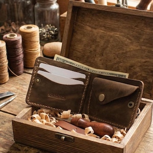 May include: A brown leather wallet, marked "Smith & Co", sits open in a wooden box. The wallet holds white cards and a folded US dollar bill. A small wooden tool and wood shavings are also in the box.