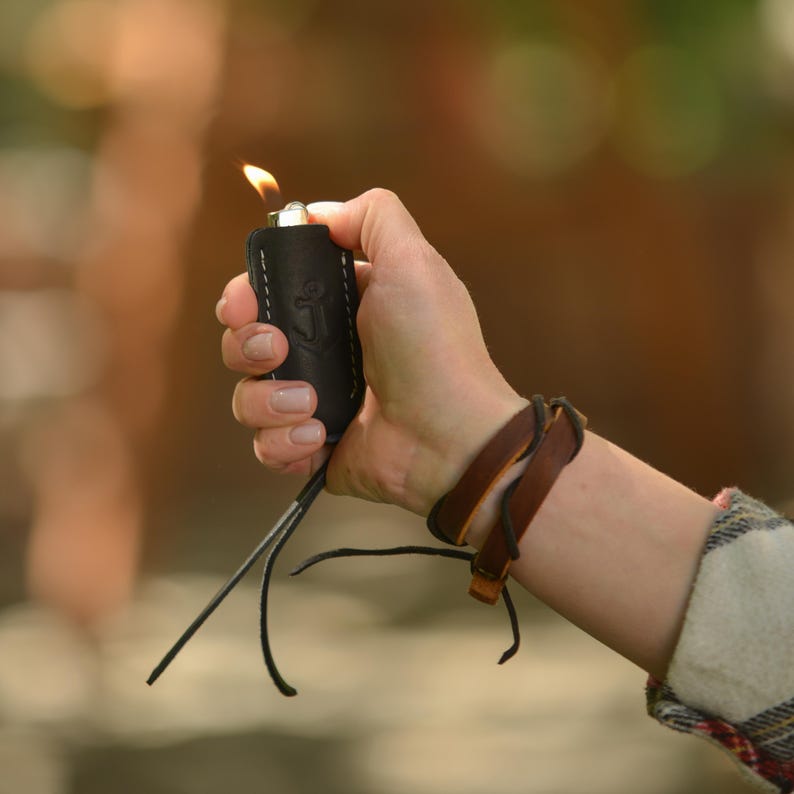 May include: A black leather lighter case with white stitching and an anchor emblem is held in a hand. The lighter is lit, with a flame visible. A brown leather bracelet is worn on the wrist.