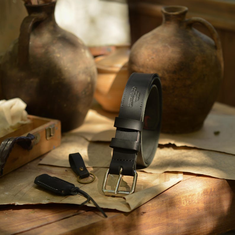 May include: A black leather belt with a silver buckle, rolled up on a wooden surface. Two black leather keychains are also visible. The belt has the text "Smith & Co" embossed on it. The background includes two brown ceramic jugs.