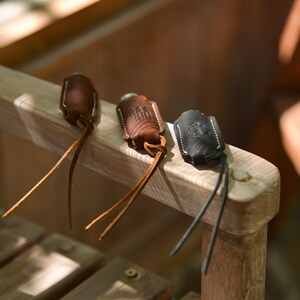 May include: Three leather keychains in varying shades of brown and black rest on a wooden surface. Each keychain features a stitched design and a leather strap. The keychains are in focus, with a blurred wooden background.