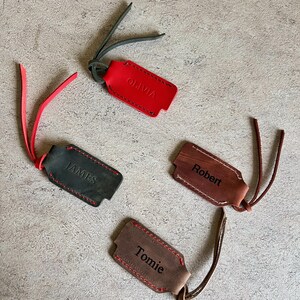 May include: Four personalized leather luggage tags in red, black, brown, and tan. Each tag has a different name: Olivia, James, Robert, and Tomie. The tags have contrasting stitching and leather straps.