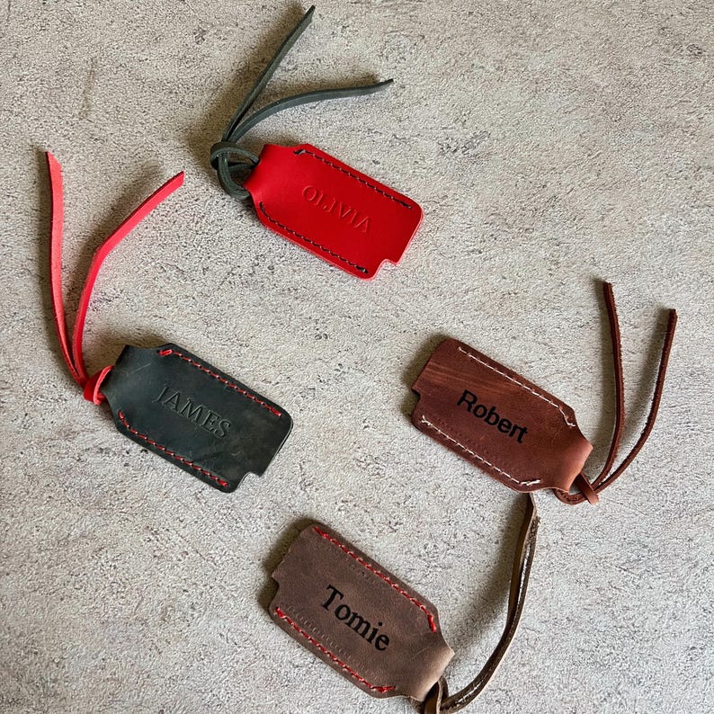 May include: Four personalized leather luggage tags in red, black, brown, and tan. Each tag has a different name: Olivia, James, Robert, and Tomie. The tags have contrasting stitching and leather straps.
