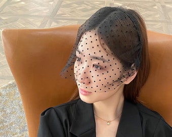 May include: A woman wearing a black polka dot veil over her hair. She is wearing a black blazer and a gold necklace with a small pendant.