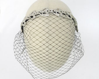 Puede incluir: Un velo de jaula de pájaros negro con una diadema de pedrería. El velo está hecho de una red fina y la diadema está decorada con un delicado diseño floral.
