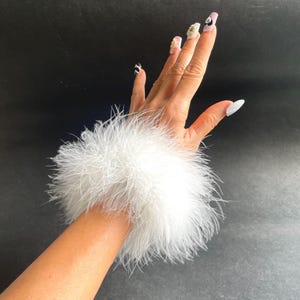 May include: A white feather wrist cuff featuring long, soft, and fluffy plumage that encircles the wrist. The accessory is constructed with a dense layer of delicate white feathers, creating a voluminous and textured appearance around the arm.