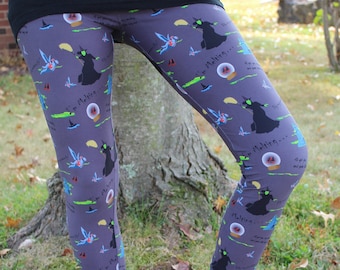 Puede incluir: Leggings grises con un patrón repetitivo de brujas en escobas, monos voladores, bolas de cristal y el texto "I'm Melting..." y "Fly my pretties". Los leggings se combinan con botines gris oscuro y una parte superior negra.