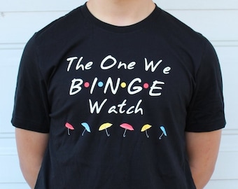 May include: Black t-shirt with the text "The One We BINGE Watch" and a row of colorful umbrellas below. 
