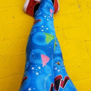 May include: Blue leggings featuring a repeating pattern of red glittery shoes, green witch legs, and white stars, paired with red glittery sneakers with white laces. The fabric includes the printed text, "There's no place like home."
