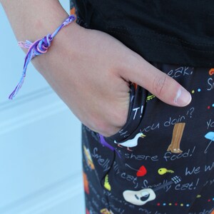 May include: A person wearing black pants with a colorful print featuring text and images of cats, birds, and other animals. The pants have a pocket on the side. The person is wearing a purple and pink friendship bracelet on their wrist.