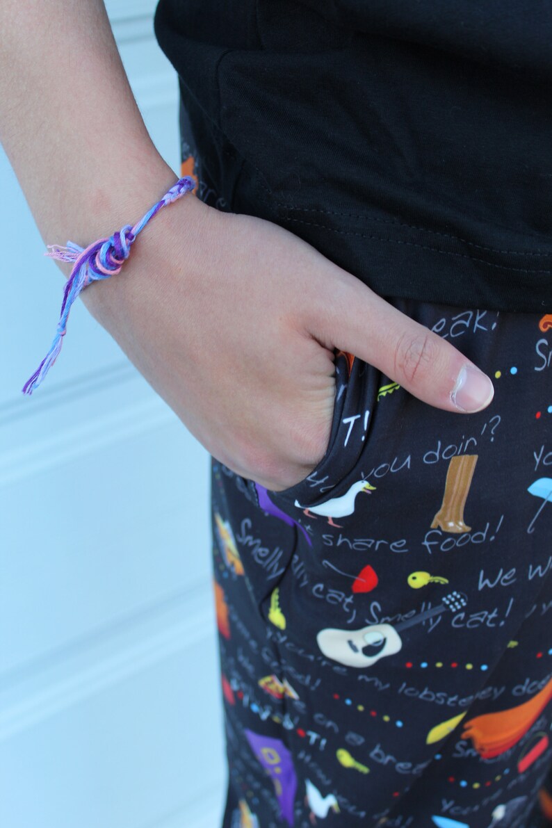 May include: A person wearing black pants with a colorful print featuring text and images of cats, birds, and other animals. The pants have a pocket on the side. The person is wearing a purple and pink friendship bracelet on their wrist.