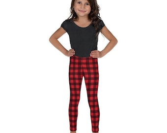 Puede incluir: Una niña con una camiseta negra y leggings a cuadros rojos y negros. Los leggings tienen un patrón de cuadros de búfalo. La niña está de pie con las manos en las caderas, sonriendo.