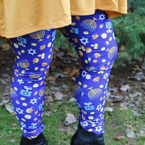 May include: A pair of blue leggings with a pattern of gold menorahs, stars of David, dreidels, and gelt. The leggings are worn with black ankle boots.