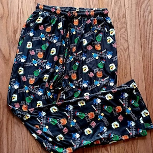 Op de afbeelding: Zwarte pyjamabroek met een herhalend patroon van kleurrijke cartoonfiguren en de tekst "PHILLY SPORTS". De broek heeft een tailleband met trekkoord en is gemaakt van een glanzend materiaal.