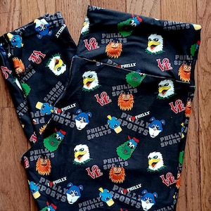 Op de afbeelding: Zwarte legging met een herhaald patroon van cartoonmascottes, het woord "PHILLY" en het woord "SPORTS" in wit en rood. Het woord "LOVE" is ook in rood te zien. De legging is op een houten oppervlak gevouwen.