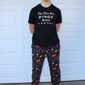 May include: A person wearing a black t-shirt with the text "The One We Binge Watch" and black pants with a colorful print of various Friends TV show icons.