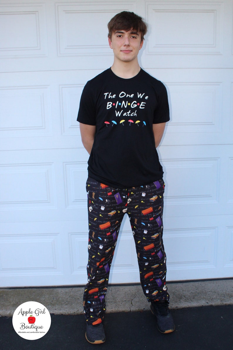 May include: A person wearing a black t-shirt with the text "The One We Binge Watch" and black pants with a colorful print of various Friends TV show icons.