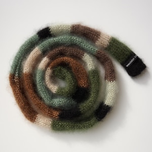 Op de afbeelding: Een opgerolde, donzige sjaal in een camouflagepatroon. De sjaal heeft blokken bruin, groen, zwart en beige. Het uiteinde van de sjaal heeft een zwart label met witte tekst. De sjaal is gemaakt van een zacht, pluizig materiaal.