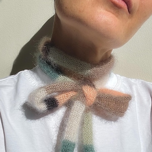 Può includere: Una sciarpa di maglia multicolore con un fiocco legato intorno al collo. La sciarpa è realizzata con un filato morbido e soffice e presenta sfumature di rosa, verde, bianco e nero.