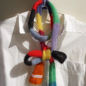 May include: A multi-coloured knitted scarf in sections of black, red, yellow, orange, blue, green, and purple. The scarf is hanging on a white hanger against a white shirt. The scarf has a tag that reads "woolcot knitwear."