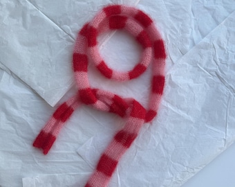 May include: A soft, striped scarf in shades of red and pink, lying on a white, textured surface. The scarf features alternating bands of solid red and pink, creating a playful and eye-catching design. The scarf is made of a fuzzy material.