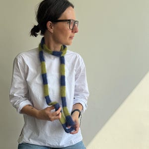 Może przedstawiać: Pasiasty szalik w odcieniach niebieskiego i zielonego. Szalik wykonany jest z miękkiego, puszystego materiału i ma prostokątny kształt. Szalik trzymany jest przez osobę ubraną w białą koszulę i niebieskie dżinsy.