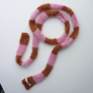 May include: A knitted scarf in pink and brown stripes. The scarf has a soft, fuzzy texture and is made up of alternating bands of light pink and a darker brown. A small label is attached to one end.