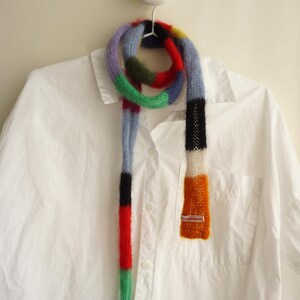 May include: A multi-coloured knitted scarf with a striped design, featuring shades of blue, red, green, and orange. The scarf is displayed on a white shirt. The scarf's dimensions are not visible.