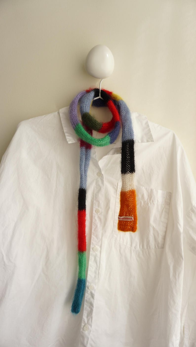 May include: A multi-coloured knitted scarf with a striped design, featuring shades of blue, red, green, and orange. The scarf is displayed on a white shirt. The scarf's dimensions are not visible.
