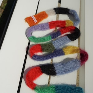 May include: A multi-coloured knitted scarf in shades of orange, red, purple, green, blue, and yellow. The scarf is displayed on a white surface. The scarf has a label that says "knitwear".