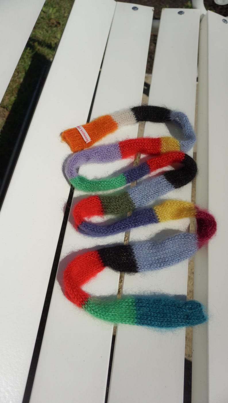 May include: A multi-coloured knitted scarf in shades of orange, red, purple, green, blue, and yellow. The scarf is displayed on a white surface. The scarf has a label that says "knitwear".