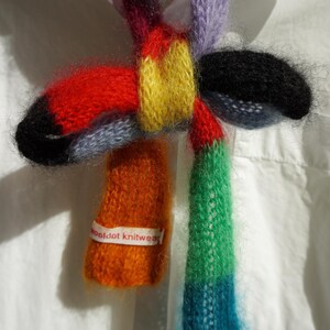 May include: A multi-coloured knitted scarf with an abstract design. The scarf incorporates red, yellow, blue, green, and orange hues. The label reads "wooldot knitwear". The scarf is displayed against a white background.