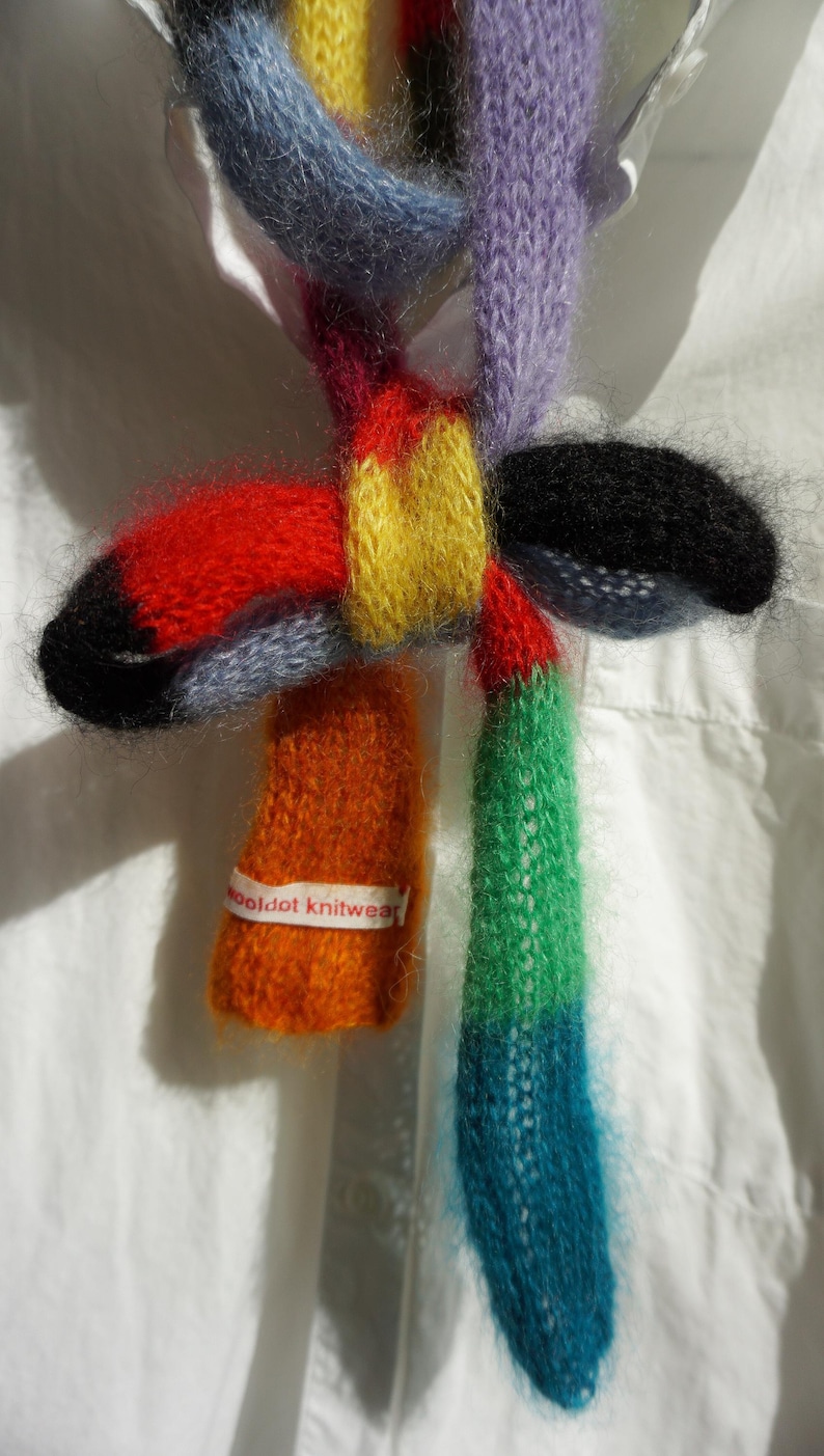 May include: A multi-coloured knitted scarf with an abstract design. The scarf incorporates red, yellow, blue, green, and orange hues. The label reads "wooldot knitwear". The scarf is displayed against a white background.