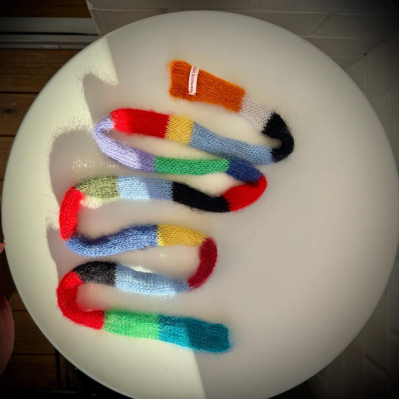 May include: A hand-knitted scarf in a winding shape, featuring segments of red, orange, yellow, green, blue, and black. A small tag with text is attached to the orange section. The scarf is displayed on a white, rounded surface.