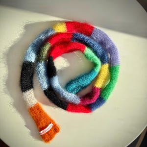 May include: A vibrant, hand-knitted scarf with a patchwork design. The scarf displays a spectrum of colours, including red, blue, green, yellow, and orange. The scarf is made of a soft, fuzzy material and is labelled "wildish knitwear".