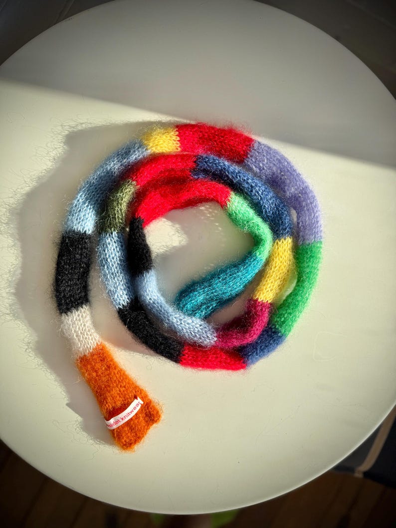 May include: A vibrant, hand-knitted scarf with a patchwork design. The scarf displays a spectrum of colours, including red, blue, green, yellow, and orange. The scarf is made of a soft, fuzzy material and is labelled "wildish knitwear".