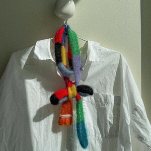 May include: A vibrant, hand-knitted scarf with a patchwork design in red, orange, yellow, black, green, and blue. The scarf is displayed on a white shirt. The label on the scarf reads "delicat knitwear".