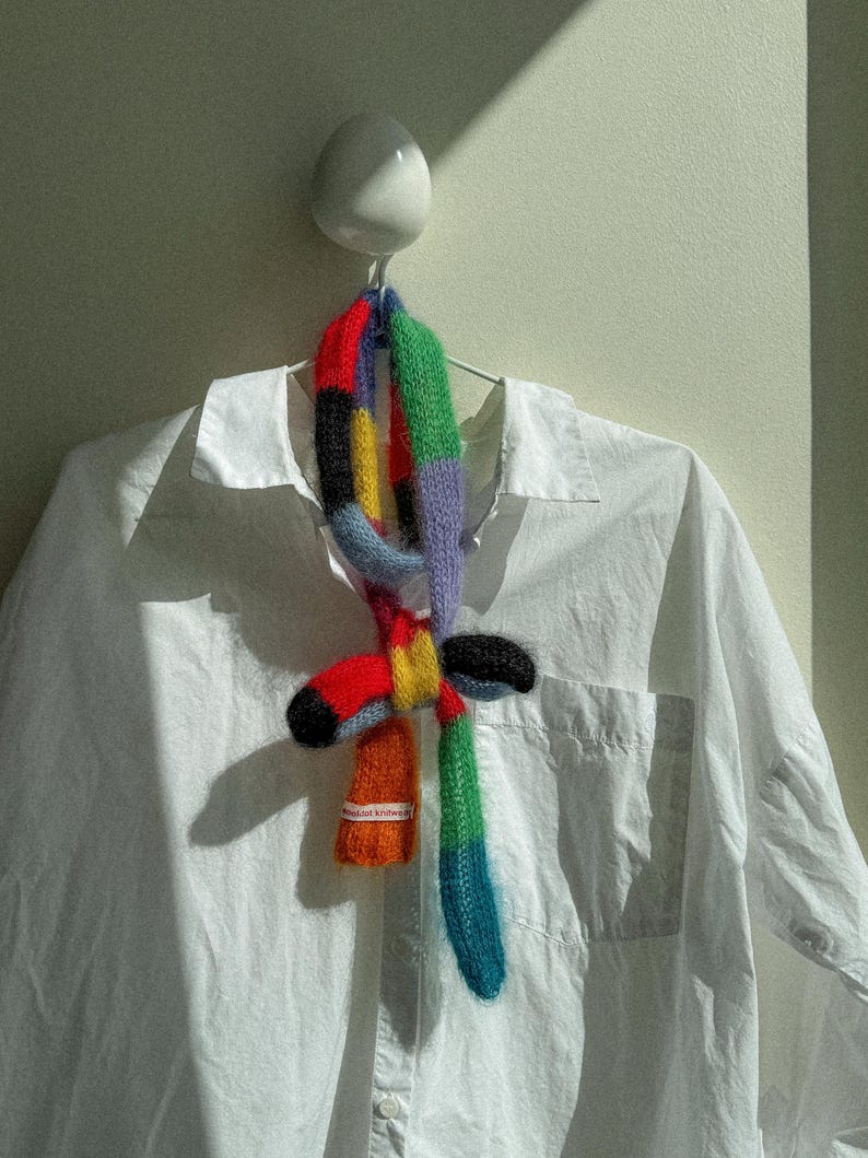 May include: A vibrant, hand-knitted scarf with a patchwork design in red, orange, yellow, black, green, and blue. The scarf is displayed on a white shirt. The label on the scarf reads "delicat knitwear".